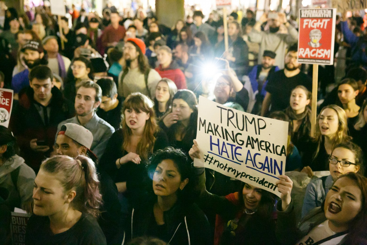 People gather to protest, holding signs about fighting racism that say that Trump is Making America Hate Again.