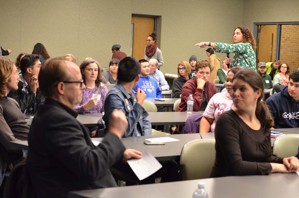 Ralina Joseph holds a microphone and points while standing the background of a workshop full of people sitting at long desks.