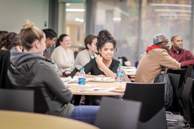 People sit at round tables with papers and water bottles while one person smailed into the camera.