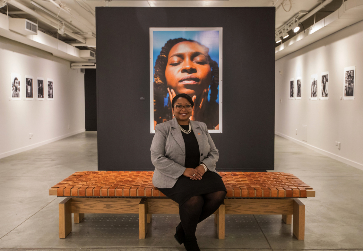 Ralina Joseph sits on a low bench in an artist gallery in front of an image of a girl with her eyes closed and her hands on her chin.