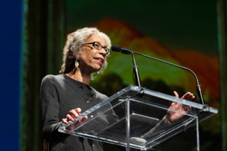 Ruth Wilson Gilmore stands behind a glass podium and speaks into a microphone.