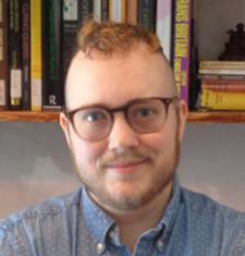 Adrian Kane wears glasses and a blue shirt with a bookcase in the background.