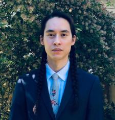 Andreas P. Bassett stands in front of a large shrubbery while wearing a dark jacket blue shirt and tie.