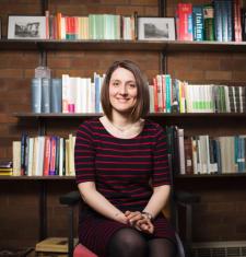 Profile of Beatrice in front of a book case. She has short brown hair and is wearing a sweater while smiling at the camera.