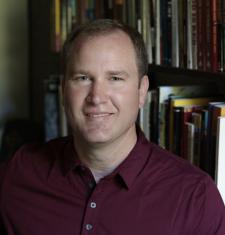 Christopher Teuton sits in front of a bookcase wearing a maroon shirt.