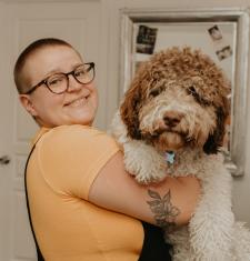 Diana Molkova stands facing the right while looking into the camera and holds a huge fuzzy dog.