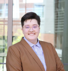 A white butch person with glasses smiling in a light brown blazer and blue and white checkered button-up shirt.