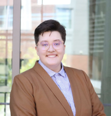 A white butch person with glasses smiling in a light brown blazer and blue and white checkered button-up shirt. 