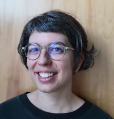 Portrait of Hannah Frydman standing in front of a brown wall wearing glasses.