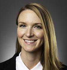 Kristin Dew stands in front of a dark wall wearing a white shirt and dark jacket.