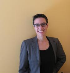 Picture of Sarah Levin-Richardson standing in front of a beige wall wearing glasses, a blazer, and a black shirt.