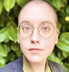 Maxine Savage stands in front of a large bush while wearing glasses and a grey jacket.