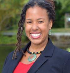 profile of Maya Smith smiling at the camera, hair pulled back with some curls along the left side, wearing a red top under a blue blazer with a necklace