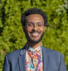 Nathanael in front of a backdrop of green foliage, smiling while wearing a blue blazer and floral shirt.