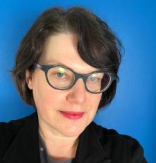 Portrait of Leigh Mercer wearing glasses and a dark jacket standing in front of a blue wall.