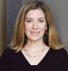 Nora smiling at the camera, shoulder length hair framing her face, wearing a black a v-neck sweater and standing in front of a gray background