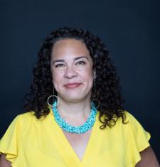 Ralina Joseph smiles in this headshot and wears a yellow flutter-sleeve blouse, a blue necklace, and long circle earrings. 