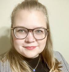 Samantha Thompson stands in front of a beige wall while wearing glasses and a jacket.