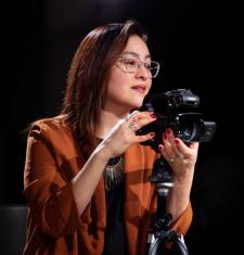 Sonia De La Cruz looks to the right while standing behind a camera on a tripod.