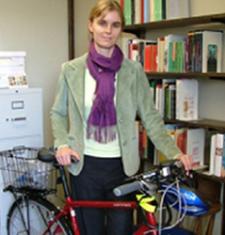 Susan Gaylard stands behind a red bicycle and in front of a bookcase wearing a light-green jacket and purple sweater.