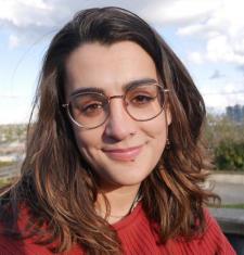 Close-up photo of a smiling woman with brown hair wearing round glasses and a red sweater. Part of the Seattle skyline is in view behind her.