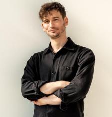 James Pierce stands in front of a white wall with his arms crossed while wearing a black collared shirt and black trousers.