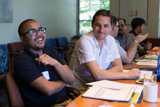 A laughing man in a black shirt sits behind a large table next to a man in a white shirt.