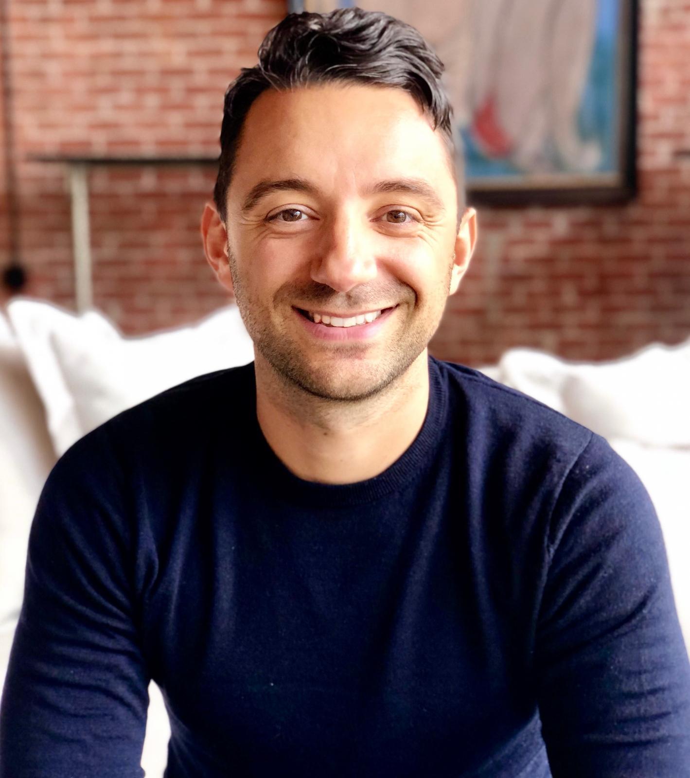 Portrait of Cristian Capotescu wearing a dark blue long sleeved shirt and sitting on a bed in front of a brick wall.