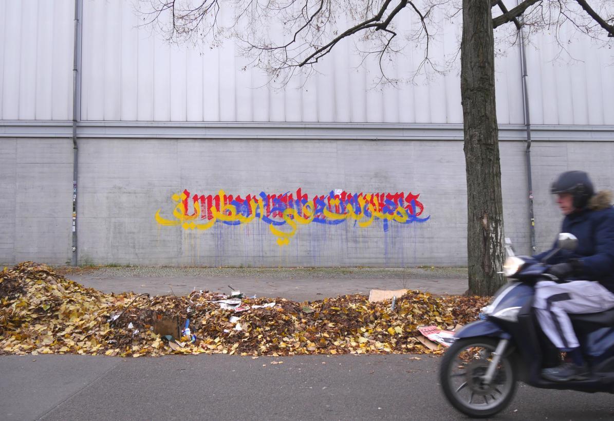 Yellow, blue, and red writing on a grey wall behind a gutter piled high with autumn leaves and street litter. On the right is a tall bare tree and an old Berliner biker zipping into the frame.