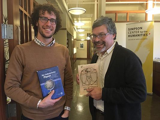 Jason Groves and Jose Alaniz each hold their books while standing in a hallway in front of a sign for the Simpson Center.