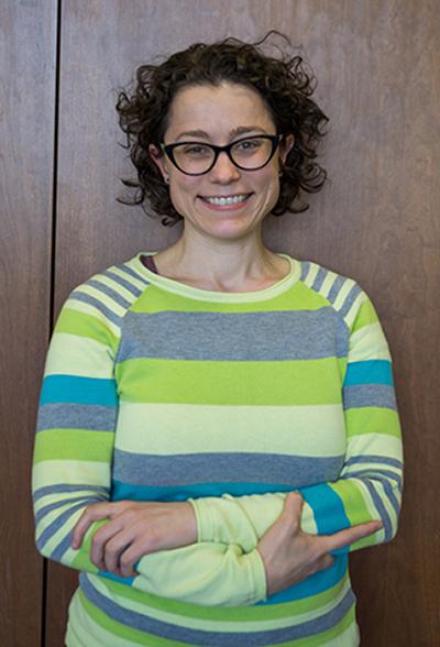 Annie Dwyer stands in front of a wood-paneled wall with arms crossed, wearing a striped green sweater.