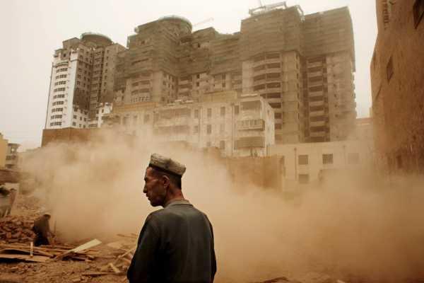 Medium shot of a Uyghur man standing in Xinjiang, with dust and buildings in the background. Photo courtesy Carolyn Drake