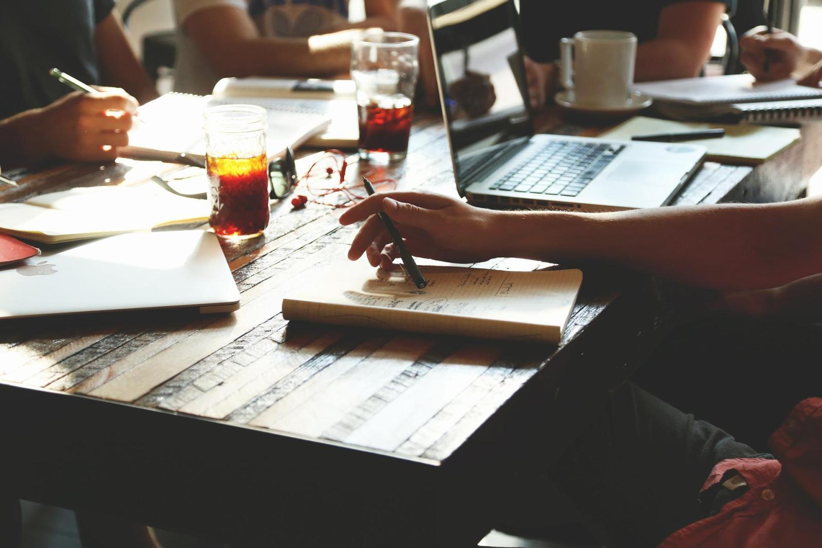 People sit around a wooden table that has notebooks, laptops, and coffee on it.