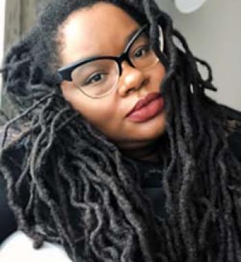 A close-up portrait of Bettina Judd wearing glasses and a dark shirt.