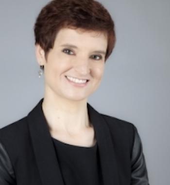 profile of Juliet Shields: short red hair, smiling at the camera, wearing a black blazer and shirt