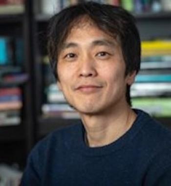 Jin-Kyu smiling at the camera in a blue sweater with blurred book stacks in the background