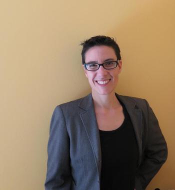 Picture of Sarah Levin-Richardson standing in front of a beige wall wearing glasses, a blazer, and a black shirt.