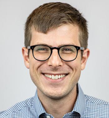 Matthew Bellinger stands in front of a white wall wearing a patterned shirt and glasses.