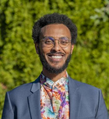 Nathanael in front of a backdrop of green foliage, smiling while wearing a blue blazer and floral shirt.