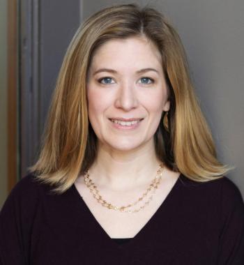 Nora smiling at the camera, shoulder length hair framing her face, wearing a black a v-neck sweater and standing in front of a gray background