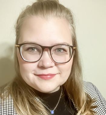 Samantha Thompson stands in front of a beige wall while wearing glasses and a jacket.