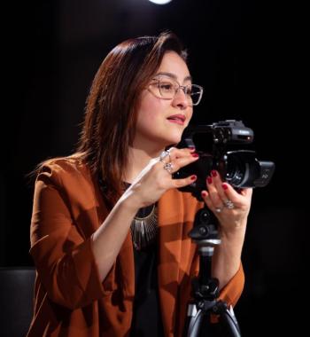 Sonia De La Cruz looks to the right while standing behind a camera on a tripod.