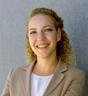 Ayda smiling at the camera in a beige blazer and white shirt, curly hair pulled halfway back and a gray background