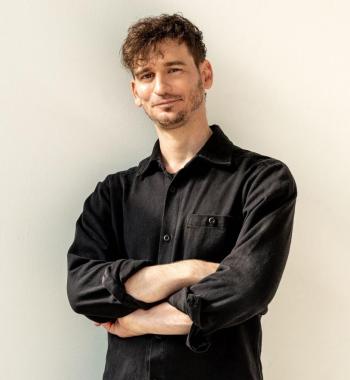 James Pierce stands in front of a white wall with his arms crossed while wearing a black collared shirt and black trousers.