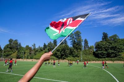 Hand holding a Kenyan flag with a scoccer field in the background
