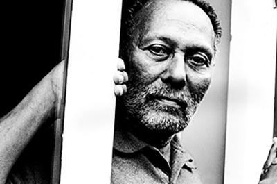 Black and white image of Stuart Hall looking at the camera from behind a pole.