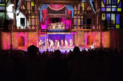 Photo of people on stage with the audience darkened in the foreground and a lit exterior of buildings in the background.