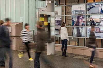 Five people walk by each other, blurred by their motion while one person is still looking at her reflection in a bank of monitors. 