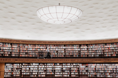 A large, circular library with two floors of bookshelves filled with colorful books. A person is pushing a cart along the upper floor walkway. The ceiling is dome-shaped and features a large, circular light fixture in the center. 