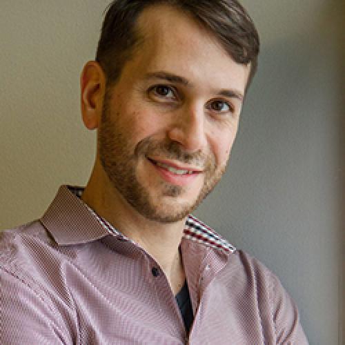 Abraham Avnisan stands in front of a white wall wearing a patterned shirt. 